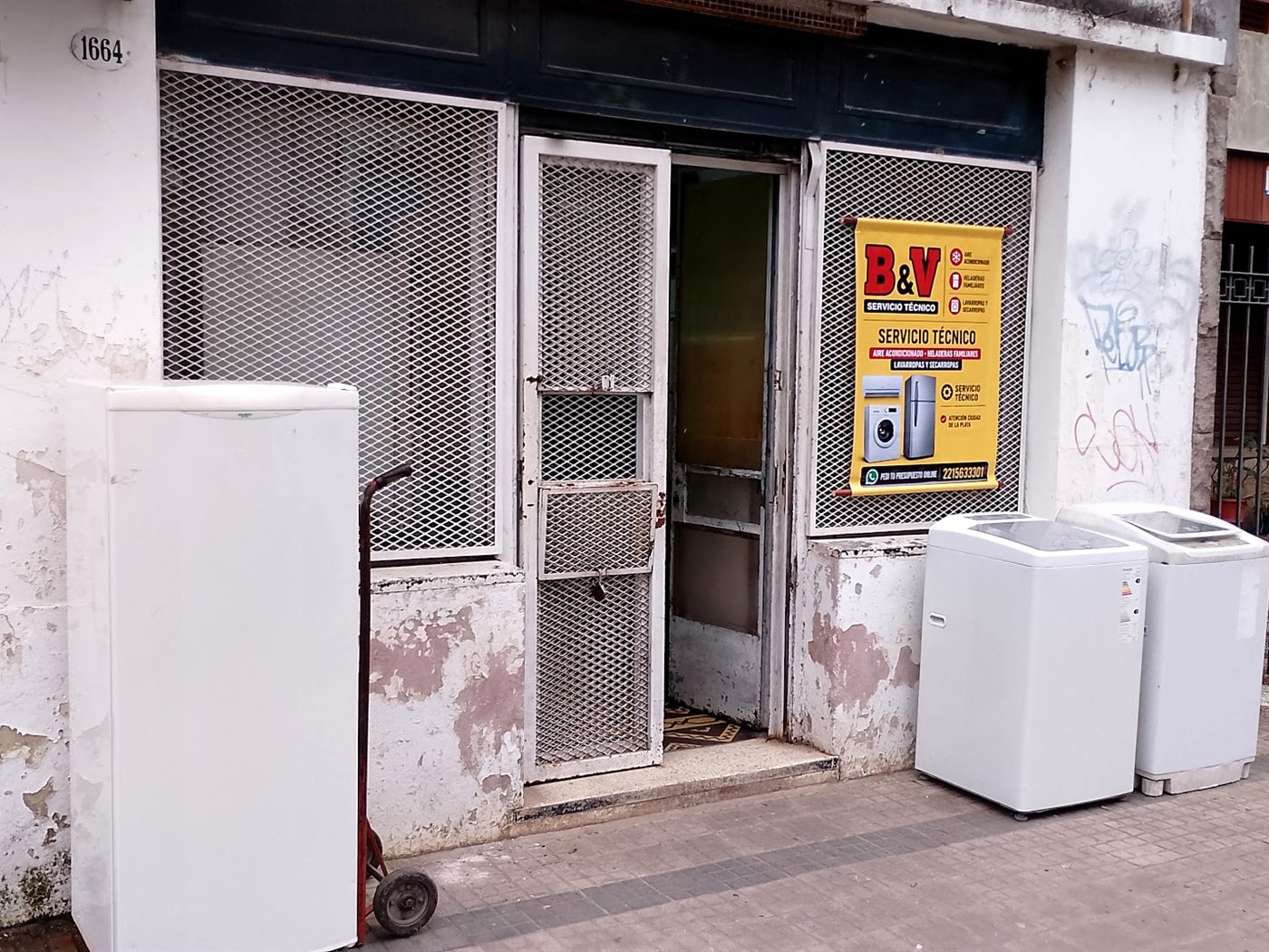 Frente del local de B&V Servicio Técnico en La Plata: el cartel de la marca sobre la reja, el numeral 1664 sobre la puerta, y equipos en la vereda esperando reparación.
